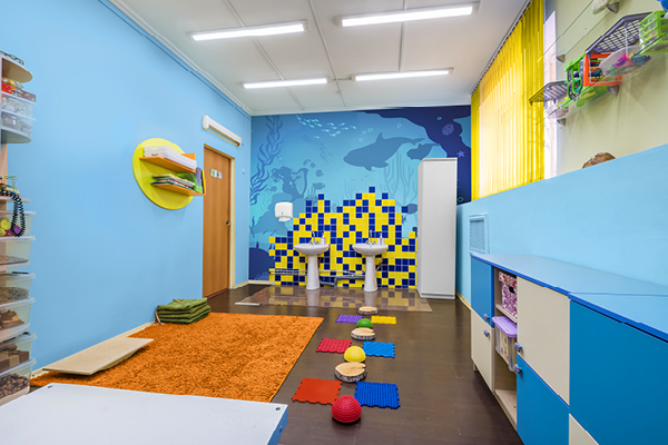 Colorful speech therapy room with blue walls, bright orange rug, sensory pathway featuring textured tiles and domes, shelves with toys, two sinks with a blue and yellow mosaic, and a large window with yellow blinds.