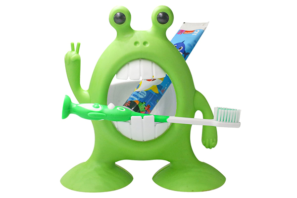 A green, cartoonish monster-shaped toothbrush holder with big eyes and a wide mouth holds a green toothbrush and a tube of toothpaste, and makes a peace sign with one hand.