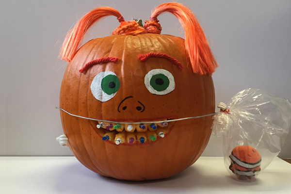 A pumpkin decorated with large green eyes, red yarn eyebrows, orange pigtails, and colorful beads for braces on its mouth. It has a plastic bag with a small pumpkin attached, resembling a trick-or-treat bag.