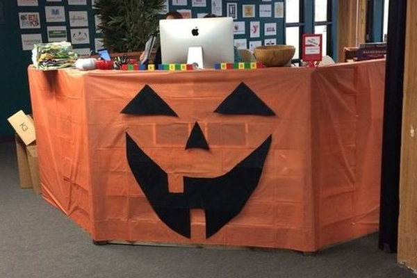A desk decorated with an orange cloth featuring a black jack-o-lantern face, creating a festive Halloween look. An iMac, folders, and office supplies sit on top of the desk.