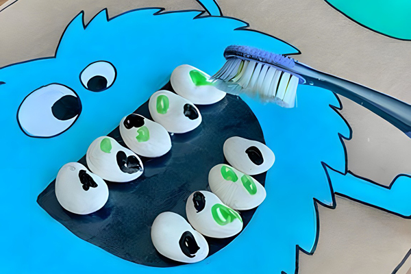A blue cartoon face with open mouth is surrounded by white oval objects with green and black marks. A toothbrush is about to brush the objects inside the mouth area.