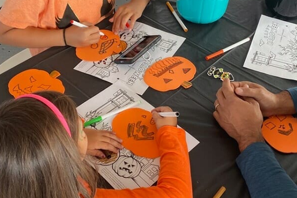 Two children and an adult decorate orange paper pumpkins with markers at a table covered in Halloween coloring pages and art supplies. A smartphone and a teal container are also on the table.