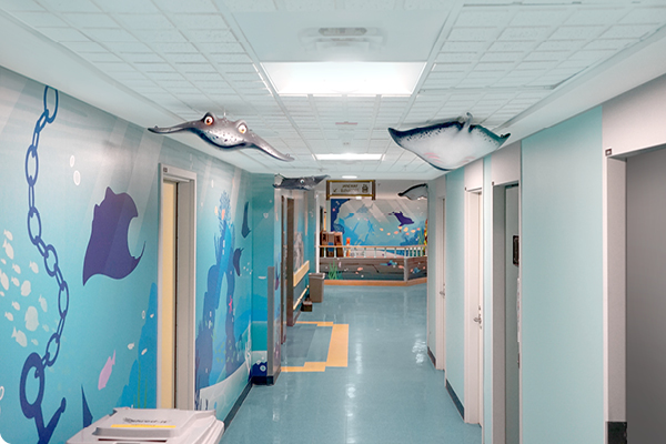 A hospital hallway decorated with ocean-themed murals and hanging manta ray models. The walls feature underwater scenes with fish and chains, creating a playful and calming atmosphere.