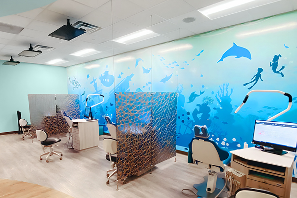 A brightly lit dental clinic with ocean-themed wall art, featuring dolphins, fish, and scuba divers. There are dental chairs and workstations separated by decorative partitions.