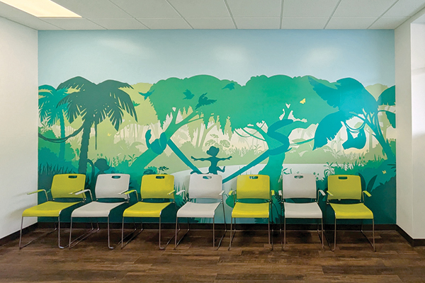 A row of green and white chairs is lined up against a wall with a mural of jungle trees, animals, and vines in shades of green and blue. The floor is dark wood, and the ceiling is white with built-in lights.