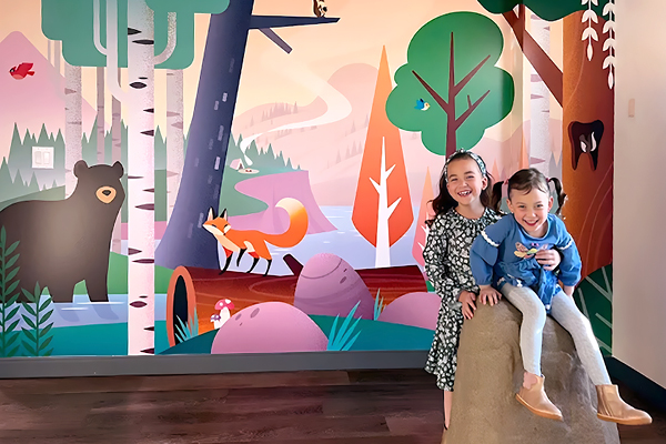 Two smiling young girls sit on a rock indoors in front of a colorful mural featuring a bear, fox, trees, and other forest scenery. The mural has a whimsical, cartoonish style.