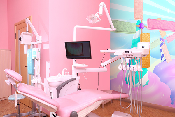 A brightly lit dental office with a pink dental chair and equipment, set against a colorful wall mural featuring whimsical windmills and pastel clouds.