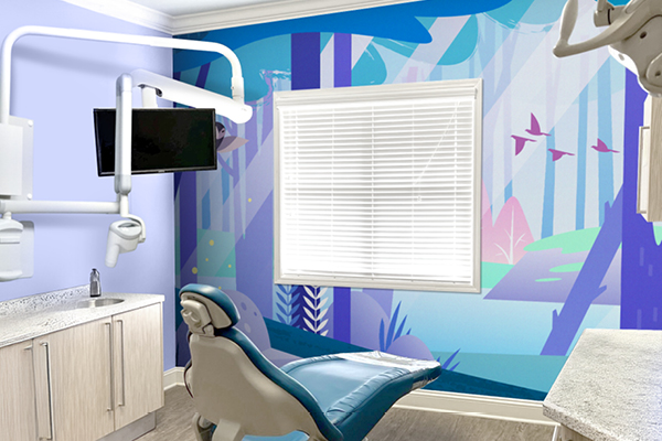 A modern dental exam room with a blue dental chair, wall-mounted equipment, and a vibrant mural of a forest with birds, in shades of blue and purple, on the wall beside a window with closed blinds.