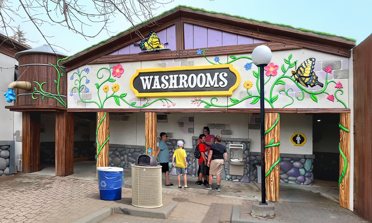 A colorful public washroom building with a large WASHROOMS sign, decorated with flowers and butterflies. Several people, including children, are gathered near the entrance and a drinking fountain.