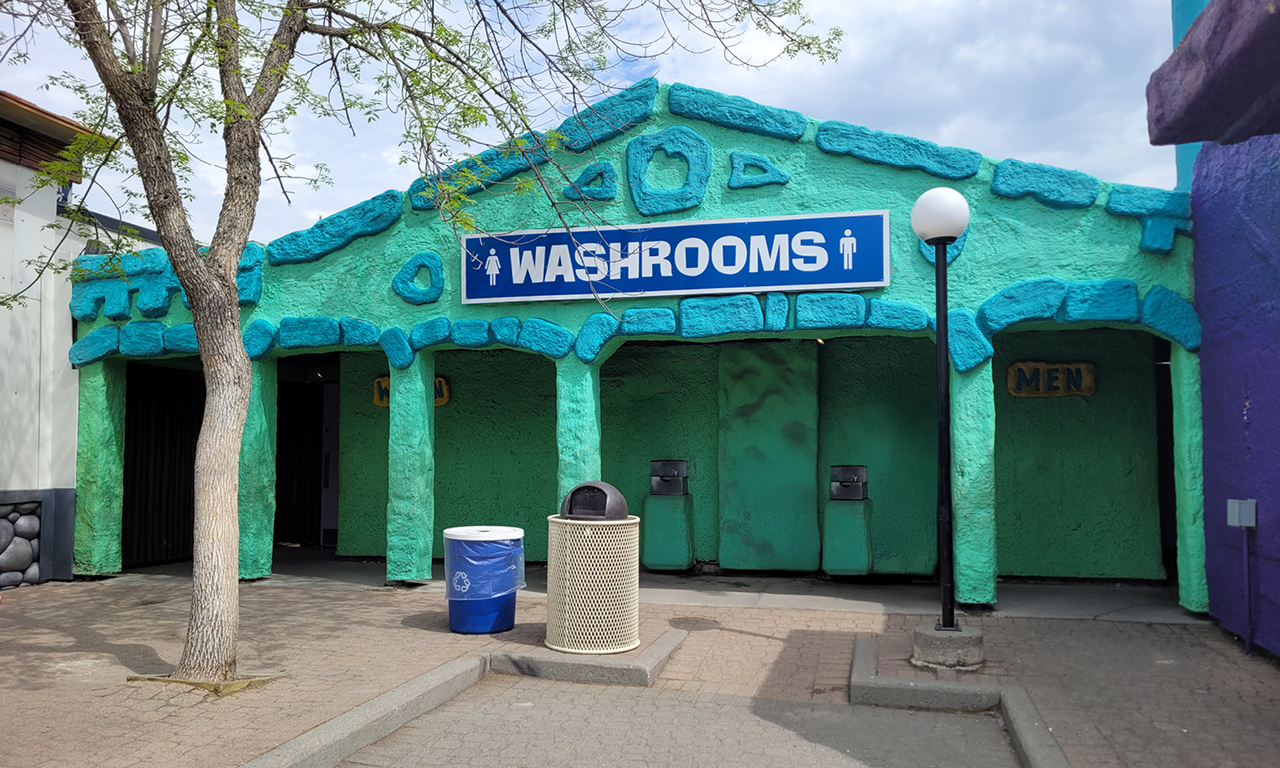 A turquoise building with a large blue WASHROOMS sign above entrances, labeled MEN and WOMEN. A tree, trash bin, recycling bin, and lamp post are in front on a paved area.