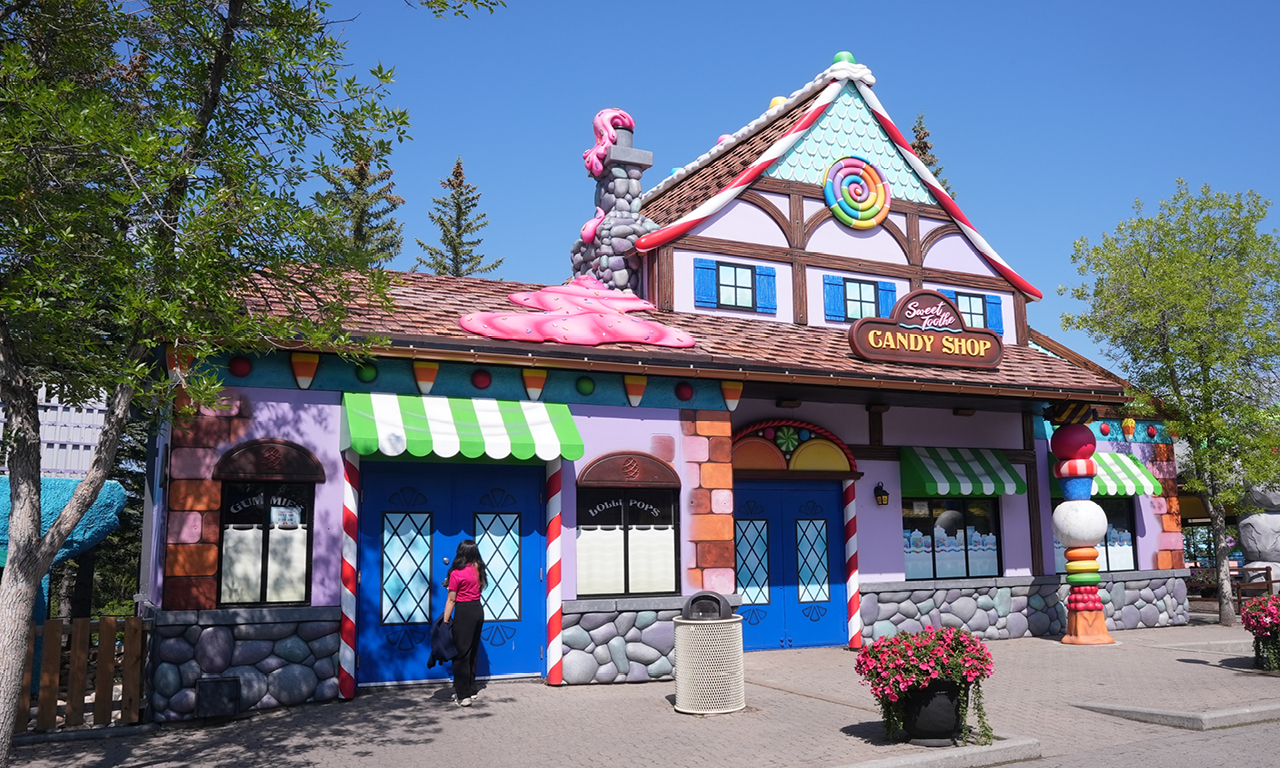A colorful, whimsical candy shop with bright striped awnings, candy decorations, and a large lollipop sign on the roof. A person stands near the entrance, and flowers are in planters outside. Trees and a blue sky are in the background.