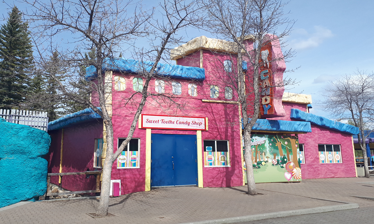 A brightly colored candy shop with a pink exterior, blue roof, and whimsical decorations. The sign reads Sweet Tooth Candy Shop. Leafless trees stand in front of the building on a sunny day.