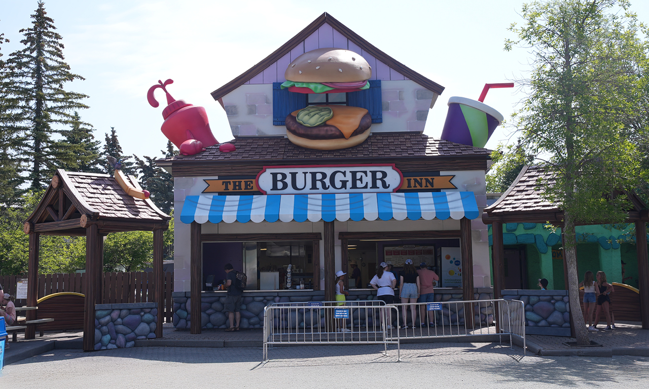 A whimsical burger restaurant features a giant burger, drink, and fries on the roof, with a blue-striped awning and people ordering at the counter. Temporary metal barriers stand in front of the entrance.