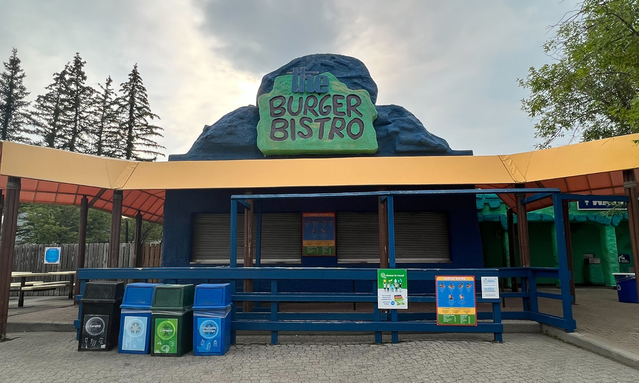A closed outdoor burger restaurant called Burger Bistro with a blue and green sign, recycling and trash bins in front, and trees in the background under a cloudy sky.