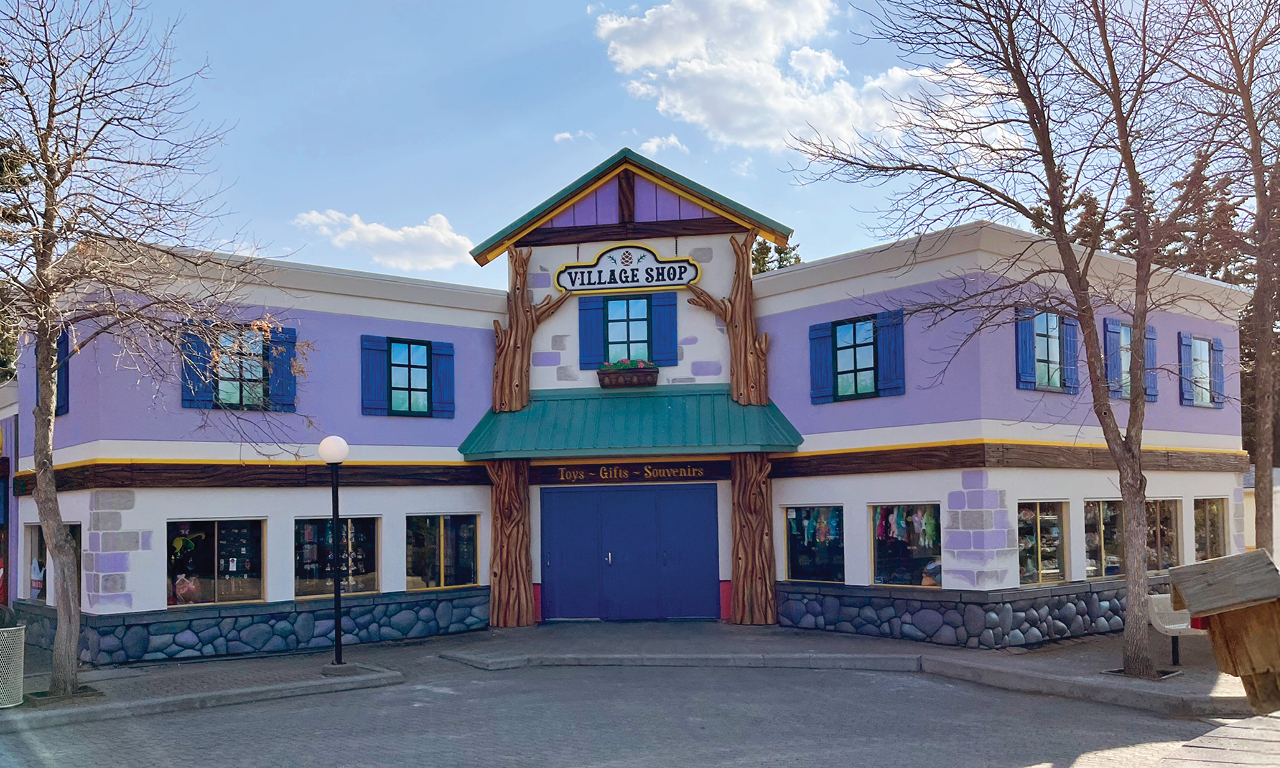 A whimsical, two-story purple building with green-trimmed windows, a green roof, and a sign reading “Village Shop.” Leafless trees stand nearby and sunlight casts shadows on the paved area in front.
