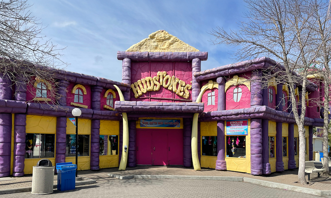 A whimsical, colorful building with bright purple and yellow walls and red trim, featuring the word KIDSTORES above double pink doors. Leafless trees and blue sky are visible, with trash bins outside the entrance.