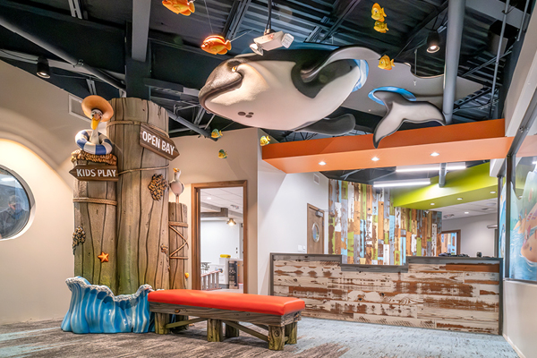 A colorful childrens waiting area with ocean-themed decor, including a large hanging orca, playful fish, a “Kids Play” wooden structure, and a red cushioned bench. The space is bright and welcoming.