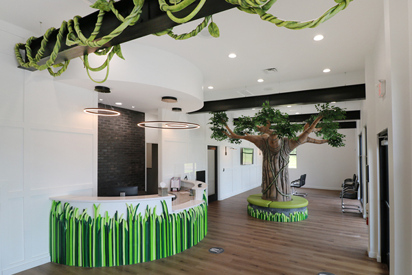 A bright, modern office waiting area featuring a round reception desk decorated with green grass designs and a large artificial tree in the center, with vines hanging from the ceiling and wood floors.
