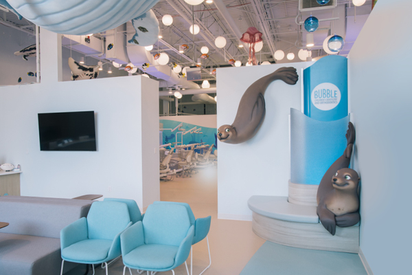 A bright, modern dental office with sea-themed decor, including two playful seal sculptures by a blue sign reading Bubble, light blue chairs, a couch, and a TV mounted on the wall.