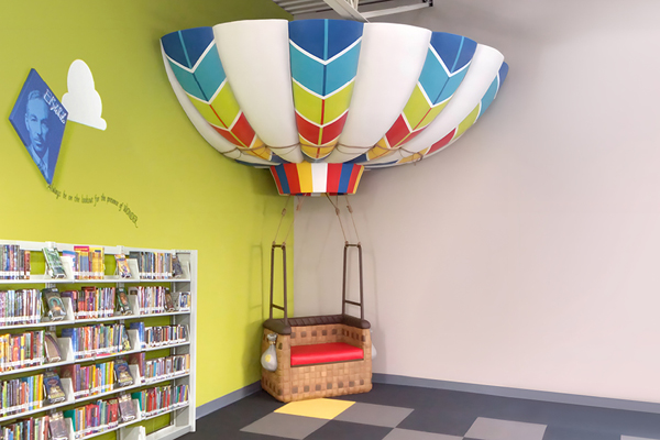 A colorful hot air balloon-themed bench is attached to a wall in a library. Nearby, shelves are filled with books, and a green wall features a cloud-shaped decoration and a quote. The floor has a checkered pattern.