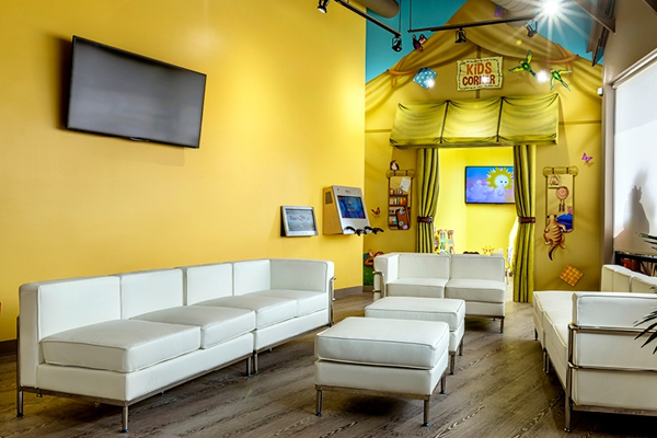 A brightly lit waiting area with white modern sofas and ottomans, yellow walls, and a colorful kids’ corner decorated with toys, books, and a TV. A larger TV is mounted on the main wall above the seating.