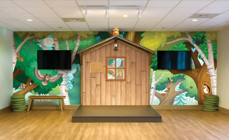 A whimsical children’s room with a wooden playhouse on a small stage, two wall-mounted TVs, forest-themed murals with cartoon animals, a bench with green cushions, and wood-style flooring.