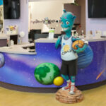 A colorful reception desk decorated with planets and space imagery, featuring a friendly blue alien statue in a basketball outfit holding a basketball, welcomes guests. The area—with purple walls and a wood-patterned floor—shows they’re hiring.