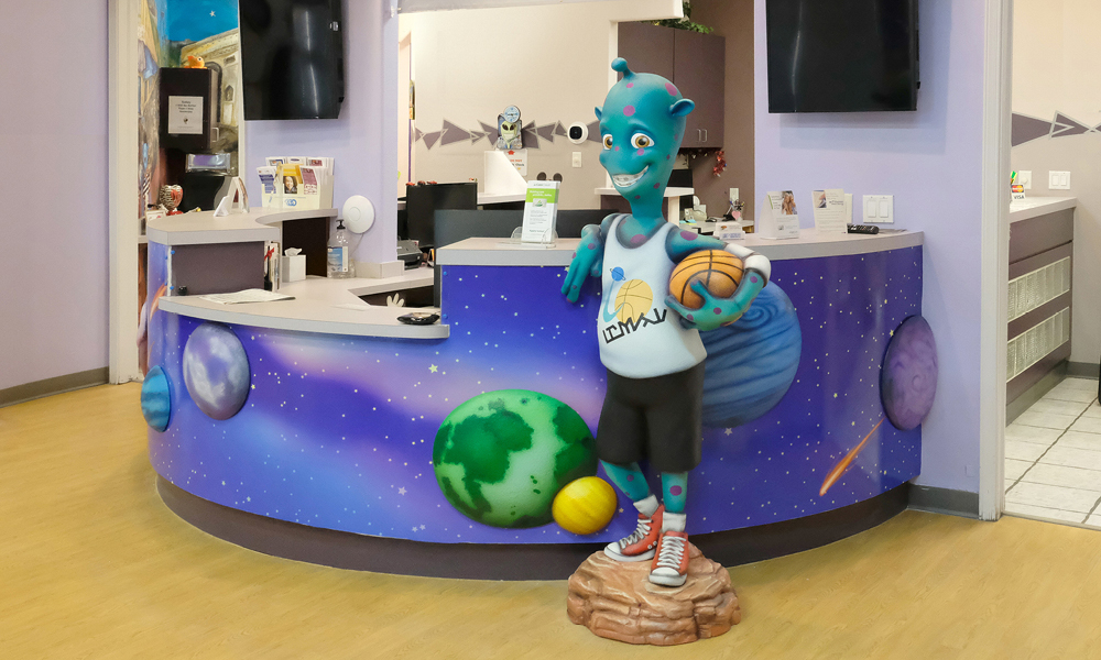 A colorful reception desk decorated with planets and space imagery, featuring a friendly blue alien statue in a basketball outfit holding a basketball, welcomes guests. The area—with purple walls and a wood-patterned floor—shows they’re hiring.