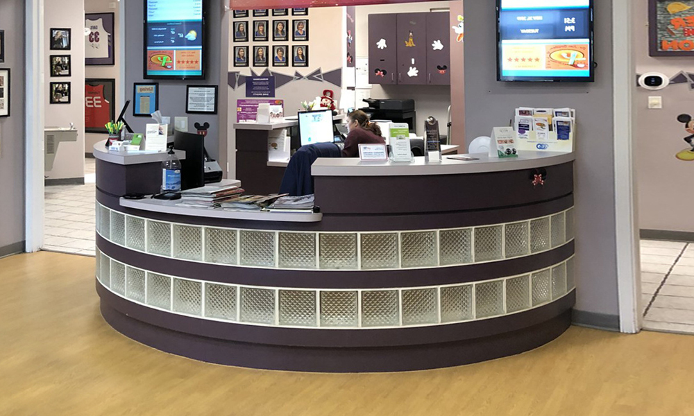 A curved reception desk in a brightly decorated office or clinic, with paperwork, a computer, and office supplies on the counter. A "hiring" sign is displayed as a person sits behind the desk and colorful decorations brighten the walls.