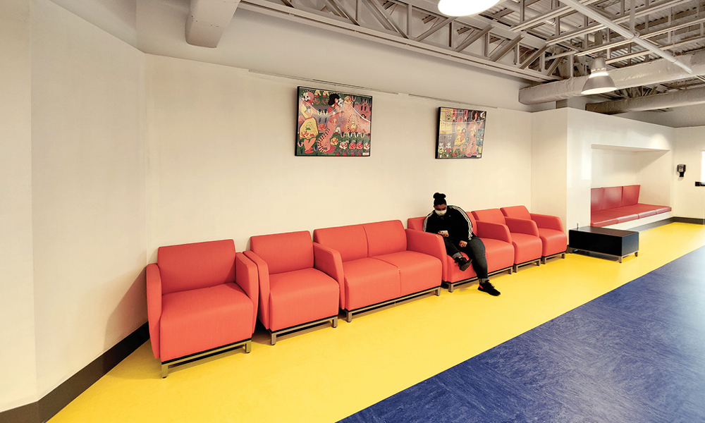 A person sits alone on a row of bright orange chairs in a spacious room, where colorful artwork and cheery flooring create a welcoming environment—perfect for those waiting during the hiring process.