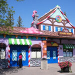 A whimsical candy shop with colorful decorations, striped awnings, candy-shaped ornaments, and a roof topped with pink icing. A person in pink stands near the blue doors beneath a "Now Hiring" sign as trees frame the scene.