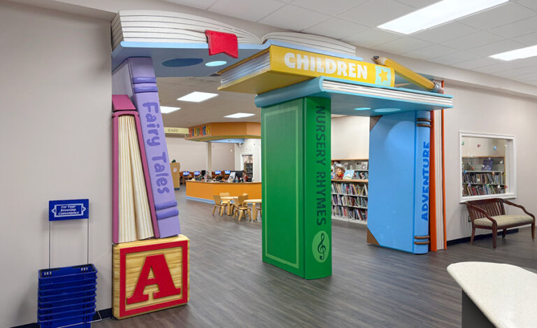 A colorful, book-themed archway with oversized books labeled “Children,” “Fairy Tales,” “Nursery Rhymes,” and “Adventure” marks the entrance to a children’s library section at Bayou Medical Clinic, filled with bookshelves and cozy reading areas.